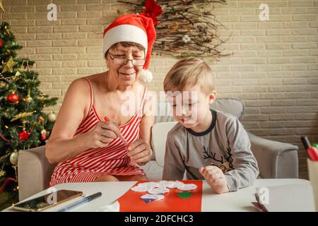 A Little boy with grandmother make Christmas craft, greeting card. Christmas tree and decoration on background. Stock Photo