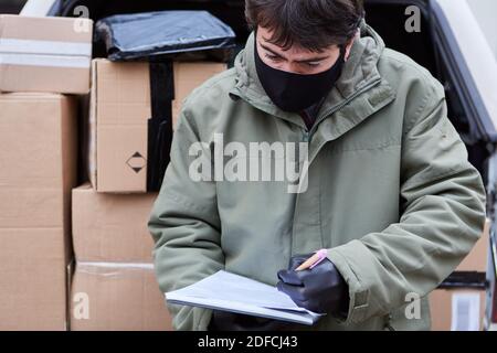 Parcel delivery man with face mask checks deliveries and stands in front of an open loading space with parcels Stock Photo