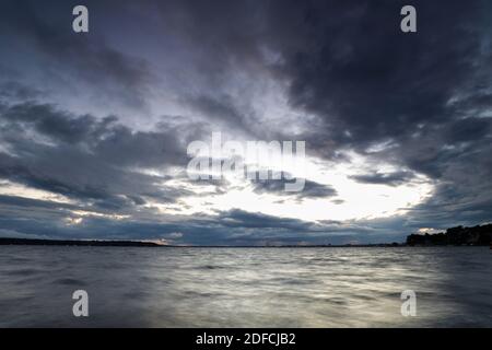Thick grey clouds dominate the sky over silvery water as the last light of the setting sun fades and nightfall approaches. Stock Photo