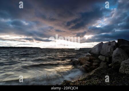 Waves crash violently onto a shoreline covered in seaweed as large rocks stand defiantly against the power of the sea.Thick storm clouds fill the sky. Stock Photo