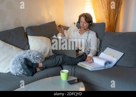 Woman, mid 50s, works from home, with laptop and communicates via headset with colleagues, home office, on the sofa, Stock Photo
