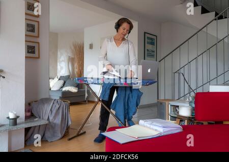 Woman, mid 50s, works from home, with laptop and communicates with colleagues via headset, home office, ironing laundry in parallel, Stock Photo