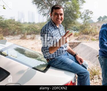 NOIDA, UTTAR PRADESH, INDIA - NOVEMBER 2020 : A young Indian man sitting on car with smile showing the victory sign. Wearing blue denim. Stock Photo