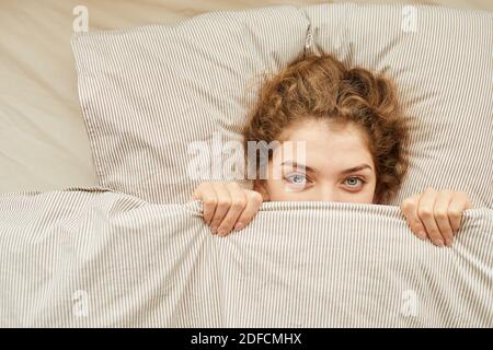 Close-up of young woman looking at camera lying on bed under the blanket Stock Photo