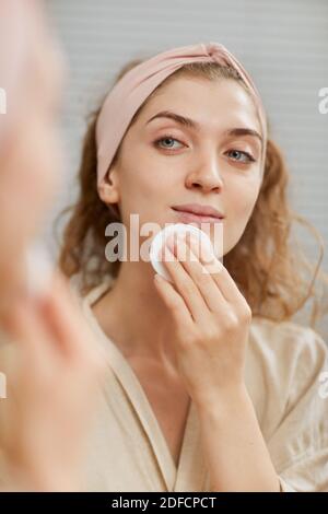Young woman looking at mirror and cleaning her face with lotion Stock Photo