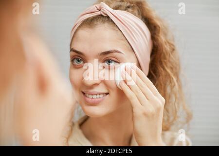 Young smiling woman looking at the mirror and removing her makeup from face with sponge Stock Photo
