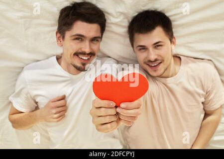 Happy couple of young gay men hide hold red paper heart lying on back out of focus on valentine's day Stock Photo