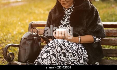 Asian woman texting on the smartphone while drinking coffee wearing fur coat and dress sits at park on old rusty bench. Resting woman in sunny day Stock Photo