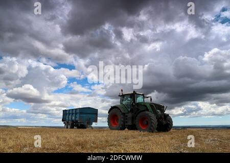 A green tractor and a trailer sit atop the recently cut remains of a field of crops under an expansive and ominous sky filled with grey rainclouds. Stock Photo