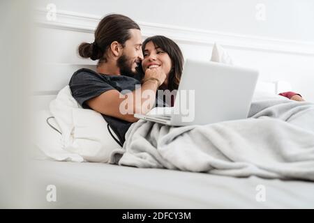 Image of romantic cheerful couple using laptop and hugging while lying in bed at home Stock Photo