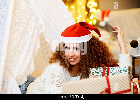 Pretty lady woman laying on Christmas giftboxes  wear knitted pullover and santa hat on bokeh lights background in decorated home room Stock Photo