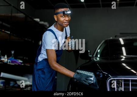 Cheerful African man worker, in uniform and protective gloves, cleaning and rubbing a car exterior by using sponge. Professional worker polishing a Stock Photo