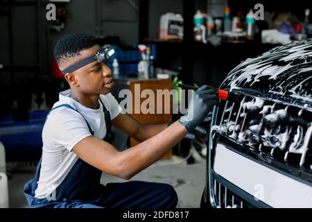 Detailing manual cleaning with soap at car wash. Young black male worker in rubber gloves washes car radiator grille of luxury black car with special Stock Photo