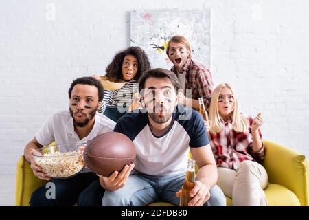 shocked multicultural friends with painted faces watching rugby championship at home Stock Photo