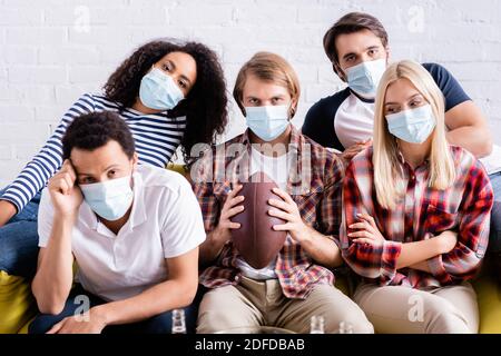 upset man holding rugby ball near sad multiethnic friends in medical masks watching competition at home Stock Photo