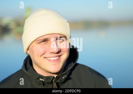 Cheerful friendly young man wearing a warm beanie hat in winter or fall looking at the camera with a beaming smile as he enjoys the sunshine outdoors Stock Photo