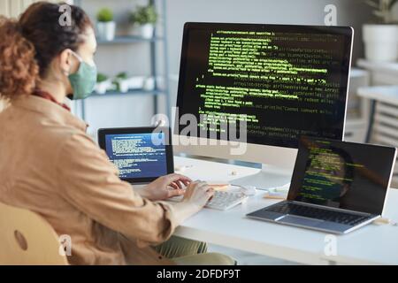 Rear view of developer in protective mask sitting at the table in front of computers and installing the program Stock Photo