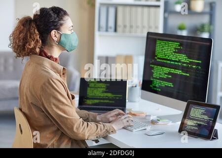 Young developer in protective mask looking at computer monitor and typing on computer keyboard working during pandemic at office Stock Photo