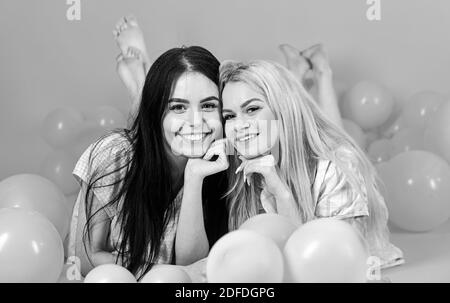 Friendship concept. Blonde and brunette on smiling faces have fun at domestic party. Girls lay on belly near balloons, pink background. Sisters or best friends in pajamas at girlish pajamas party. Stock Photo