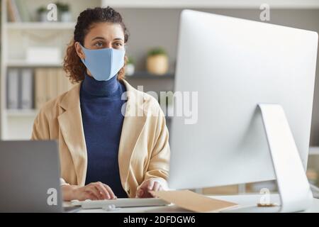 Young businesswoman in protective mask sitting at the table and working on computer she working at office during pandemic Stock Photo