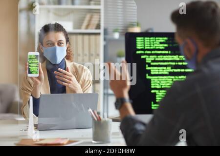 Woman in mask sitting at the table opposite her colleague and showing him mobile phone with software while working at office Stock Photo