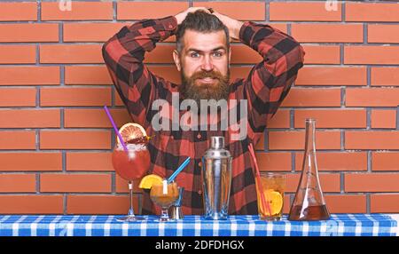 Alcoholic drinks concept. Man in checkered shirt on brick wall background prepares drinks. Barman with long beard and mustache and stylish hair on confused face forgot how to make alcoholic cocktail. Stock Photo