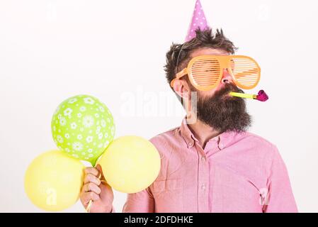 Man with beard and mustache on calm face holds air balloons, white background. Guy in party hat with party horn celebrates. Party concept. Hipster in giant sunglasses celebrating birthday. Stock Photo