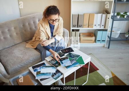Young woman in eyeglasses sitting on sofa at the table and choosing the best photos for design project Stock Photo