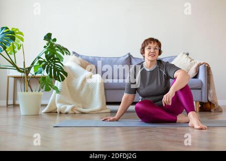 An elderly woman sits on the floor before a Pilates workout. She is looking at the camera and smiling. Helathy lifestyle concept. Stock Photo
