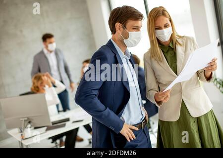 Young man and woman with protective facial masks discussing with paper in hands indoors in the office with young people works behind them Stock Photo