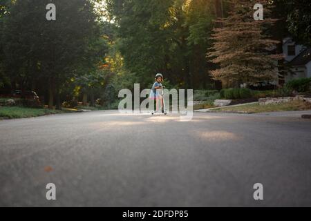 A small child rides on a scooter on  a tree-lined street in sunlight Stock Photo
