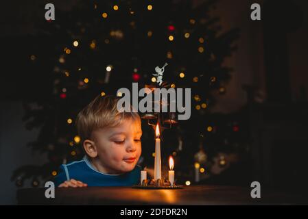 Adorable young child blowing candles with lit up christmas tree behind Stock Photo