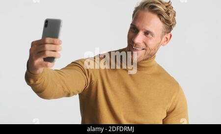 Attractive smiling bearded man wear turteneck taking selfie on smartphone over white background. Modern technology Stock Photo