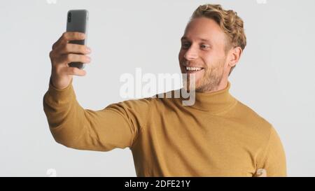 Attractive smiling bearded man in turtleneck having video call with friends using smartphone over white background. Video chat Stock Photo