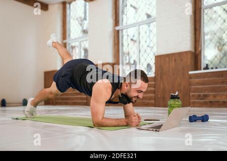 Handsome young sport man doing one leg up push ups exercise in empty gym or home watching online sports videos all in black clothes. Muscular Stock Photo