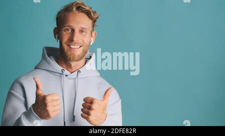 Attractive smiling bearded man in hoodie wear wireless earphones keep thumbs up near place for advertisement or promotional text isolated on blue Stock Photo