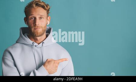 Handsome stylish bearded man in hoodie wear wireless earphones showing place for advertisement or promotional text over colorful background Stock Photo