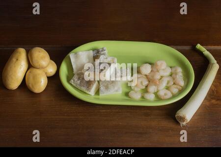 A wooden table with potatoes, leek, and a green platter with cod fillets and prawns. Ingredients for cooking fish and vegetables. Stock Photo