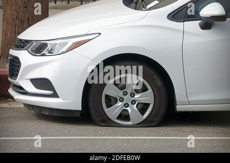 A white car with a flat tire on the front, driver side is shown parked along a city street during the day. Stock Photo