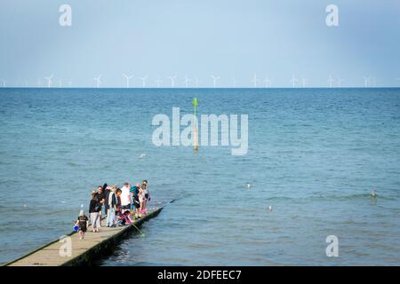 Group of people stood at the end of the jetty with wind turbines visible out at sea in the distance. Stock Photo