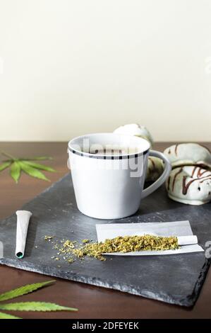 Fresh coffee, marijuana leaves, joints and chocolate donuts on wooden table with copy space top. Stock Photo