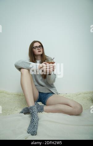 sad young woman in eyeglasses sitting on white background with mug of tea looks at camera Stock Photo