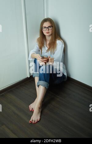sad young woman in eyeglasses sitting on floor with cup of tea looks at camera Stock Photo