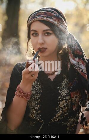 young gypsy woman smoking pipe in autumn forest, looking at camera