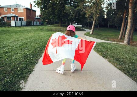 Girl in sport helmet wrapped in large Canadian flag riding on roller skates in park. Canada Day celebration outdoor. Kid in large Canadian flag Stock Photo