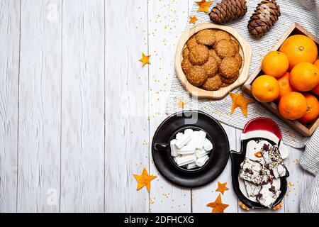 Cozy winter still life with tangerines, cookies, chocolate, pine cones and a cup of marshmallows on a light wooden table. Top view, place for text. Stock Photo