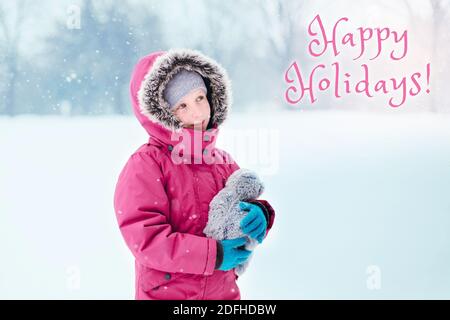 Happy holidays. Christmas card with text. Caucasian smiling girl child looking at  falling snow on cold winter snowy day outdoor. Traditional Christma Stock Photo