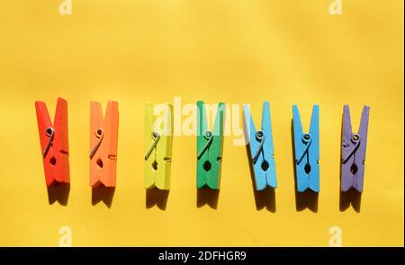Seven wooden multi-colored clothespins, painted in the colors of the rainbow, on a yellow paper background, lie in a row. Stock Photo