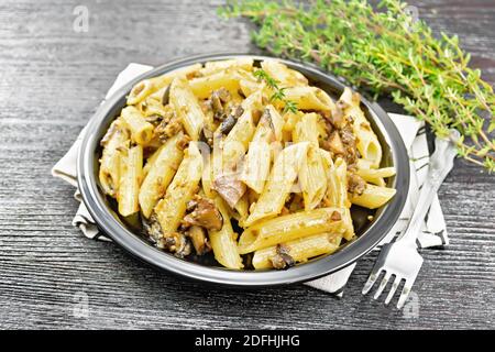 Penne pasta with wild mushrooms in a plate on a towel, thyme, fork and garlic on a wooden board background Stock Photo