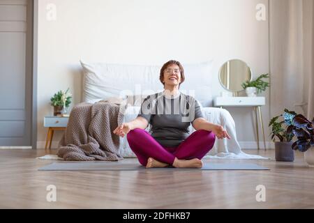 An elderly woman meditates at home in the lotus position. The concept of a healthy and active lifestyle in old age. Stock Photo